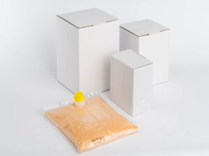 Unsere Produkte 4 Ein Foto, das drei weiße Kartons unterschiedlicher Größe und einen versiegelten transparenten Plastikbeutel mit einer orangefarbenen Flüssigkeit und einer gelben Ausgießeröffnung zeigt, der vor den Kartons auf einer weißen Oberfläche platziert ist und an ordentlich gestapelte Eierkartons erinnert.