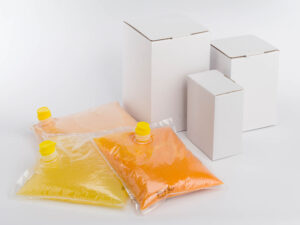 Unsere Produkte 2 Im Vordergrund sind drei Plastikbeutel mit Flüssigkeiten in den Farben Gelb, Orange und Hellrosa zu sehen, jeder mit einer gelben Tülle. Dahinter sind drei weiße Kisten unterschiedlicher Größe zu sehen. Der Hintergrund ist schlicht und weiß und erinnert an eine ausgestellte Ei-Sammlung.