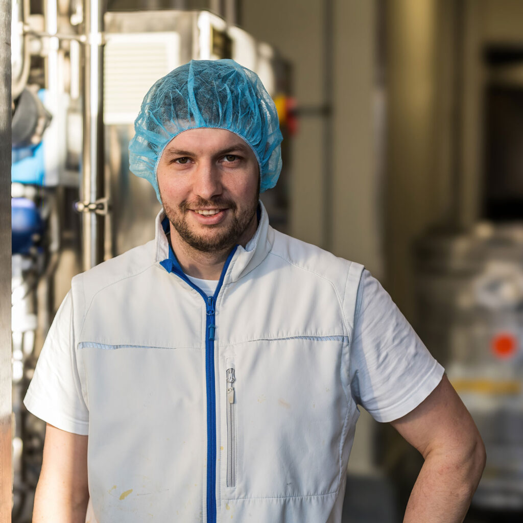 Ein Mann in einem weißen Laborkittel und einem blauen Haarnetz steht in einer Industrieanlage. Er scheint sich in einem Labor oder einer Produktionsumgebung zu befinden, mit Maschinen und Geräten im Hintergrund. Er lächelt und hat die Hände in die Hüften gestemmt.
