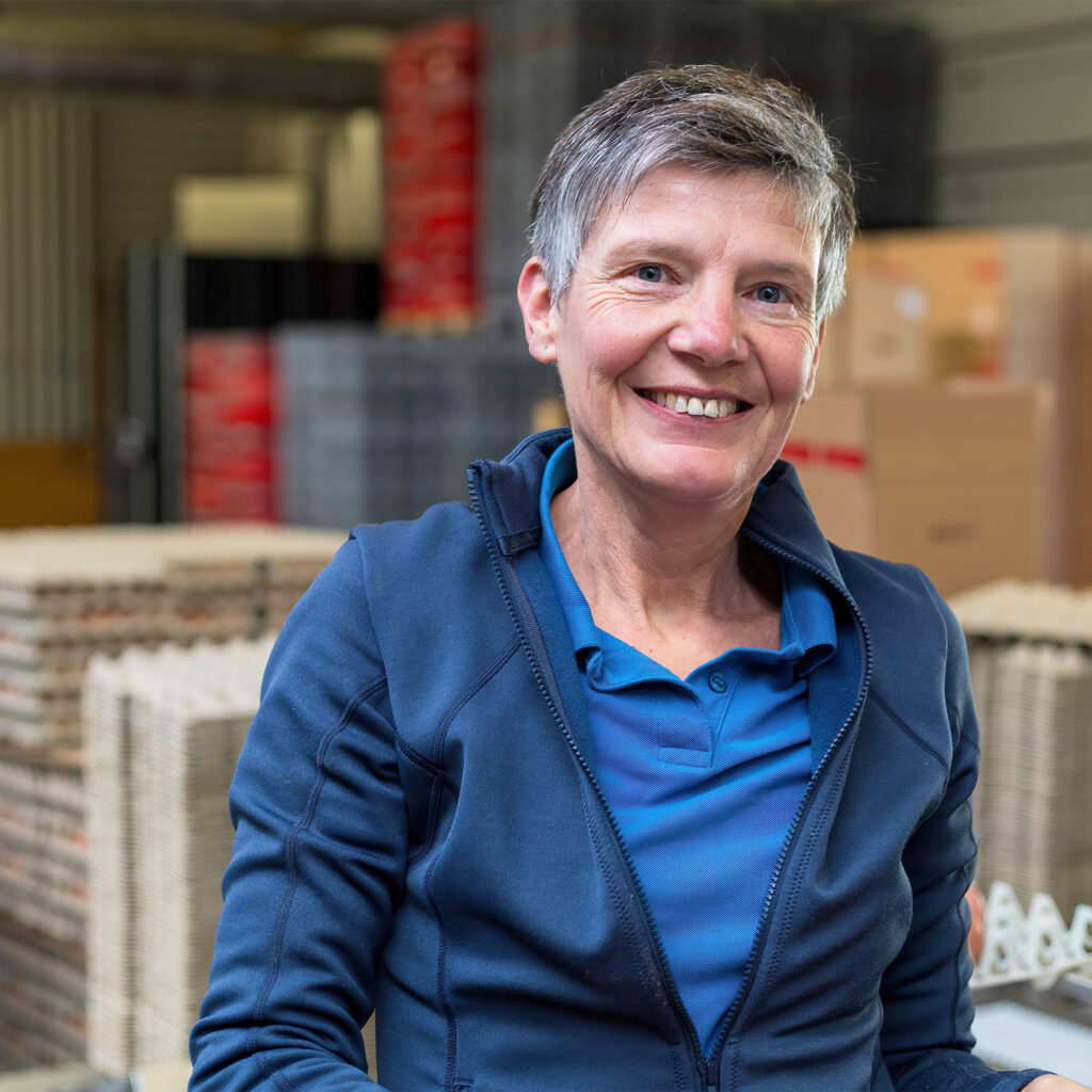 Eine lächelnde Person mit kurzen grauen Haaren, die ein blaues Poloshirt und eine dazu passende Jacke trägt, steht in einem gut organisierten Lager. Im Hintergrund sind Stapel von Kartons und Kisten zu sehen, neben anderen Industriematerialien und Zubehör von Eiers. Die Umgebung wirkt ordentlich und gut beleuchtet.