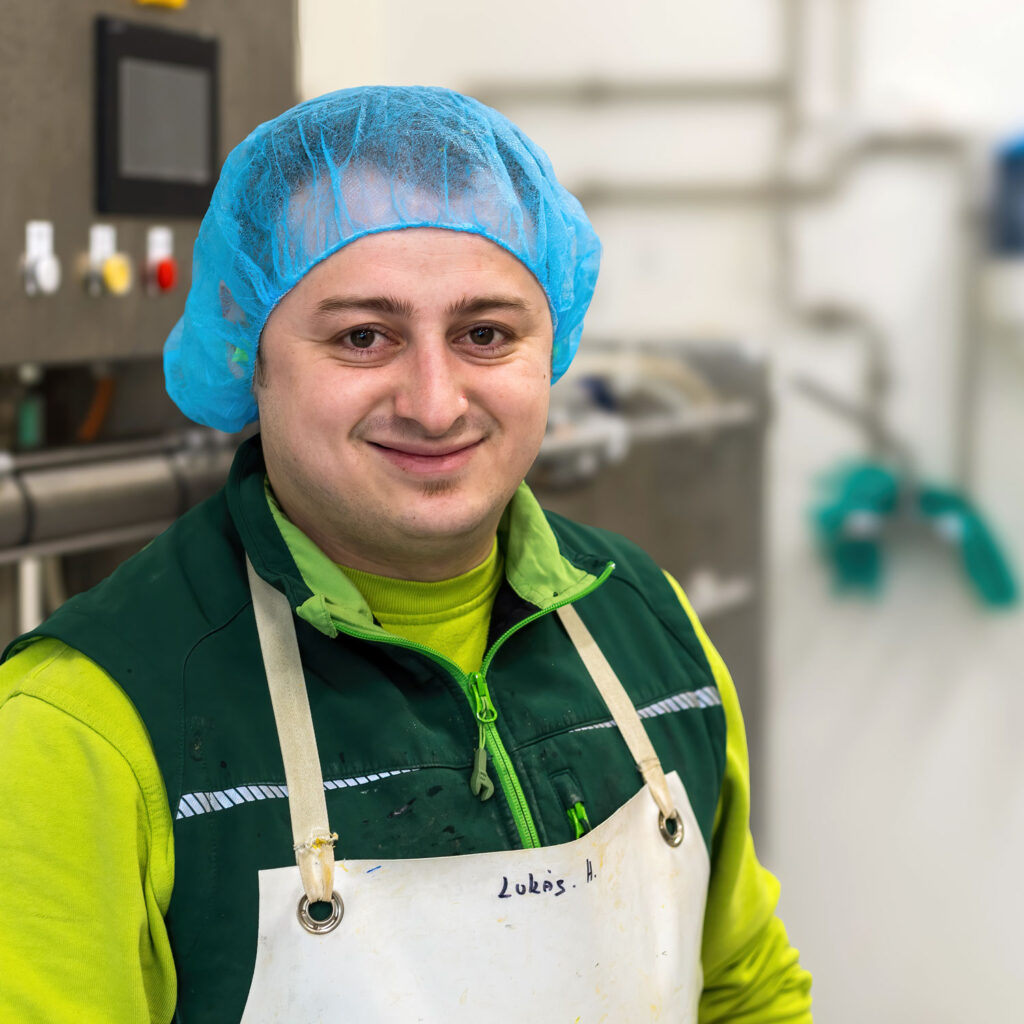 Ein Mann mit blauem Haarnetz, grüner Jacke und weißer Schürze steht in einer Arbeitsumgebung mit Industrieanlagen im Hintergrund. Er lächelt und scheint sich in einer Eierverarbeitungsanlage zu befinden. Der Name auf seiner Schürze lautet „Lukas“.