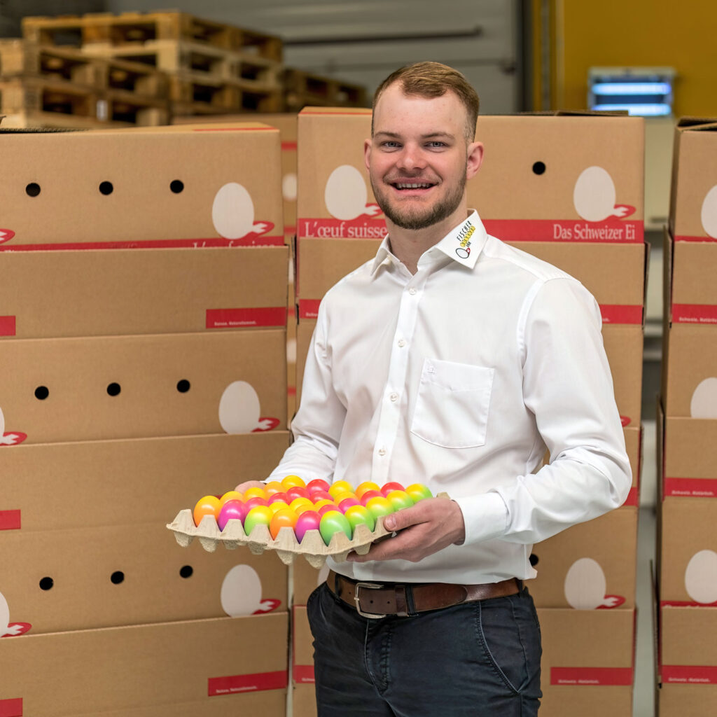 Ein Mann in einem weißen Hemd mit Kragen hält einen Karton mit bunten Eiern. Er steht vor gestapelten Kartons in einem Lagerhaus. Die Kartons sind mit Bildern von Eiern beschriftet. Der Mann lächelt und scheint an der Eierproduktion oder -verteilung beteiligt zu sein.