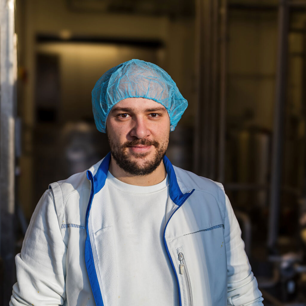 Ein Mann in weißer Jacke und blauem Haarnetz steht in einer Industrie- oder Laborumgebung und verarbeitet möglicherweise Eis. Er hat einen Bart und blickt mit neutralem Gesichtsausdruck in die Kamera. Der Hintergrund ist verschwommen und metallische Geräte sind zu sehen.