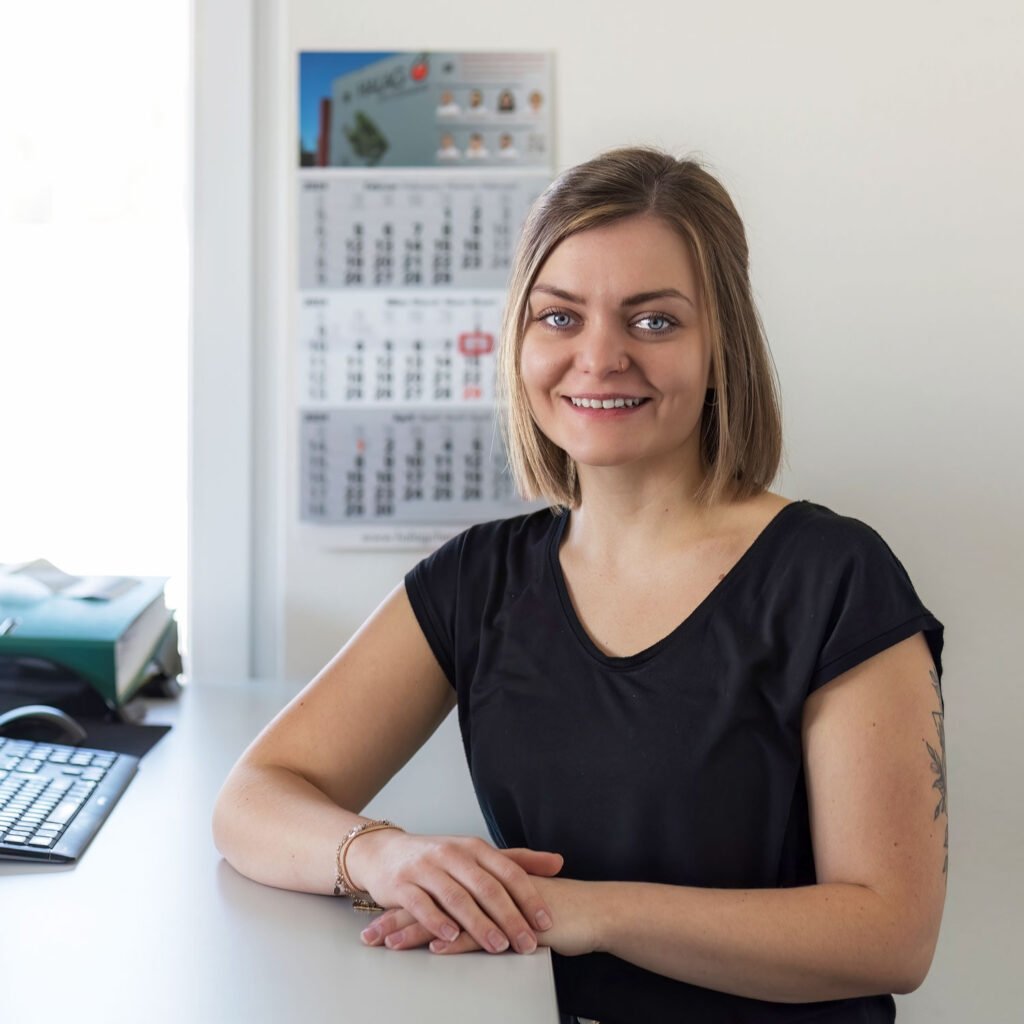 Eine lächelnde Frau mit schulterlangem Haar sitzt an einem Schreibtisch. Sie trägt ein schwarzes Hemd und ihre Hände ruhen neben einem Eisbecher auf dem Schreibtisch. Hinter ihr hängt ein Wandkalender mit mehreren Seiten und auf dem Schreibtisch ist eine Computertastatur zu sehen.
