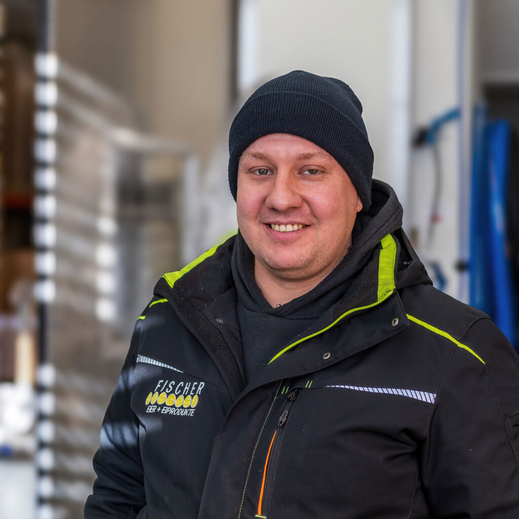 Ein Mann mit einer schwarzen Beanie-Mütze und einer schwarzen Jacke mit grünen Akzenten steht lächelnd da. Auf der linken Seite der Jacke steht der Text „FISSCHER“. Hinter ihm ist eine Industrieanlage mit einem teilweise geöffneten Garagentor und verschiedenen Geräten, darunter Stapel von Eierkisten, zu sehen.