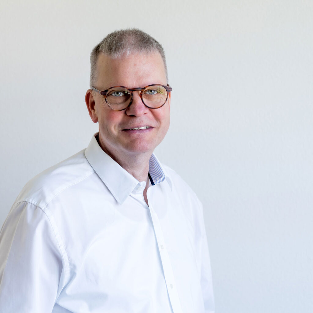 Vor einem hellen Hintergrund steht ein Mann mit kurzen grauen Haaren und Brille. Er trägt ein weißes Hemd mit Knöpfen und sein subtiles Lächeln verleiht ihm eine Aura stillen Selbstvertrauens.