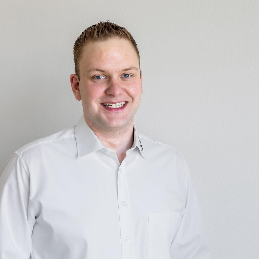 Ein junger Mann mit kurzen hellen Haaren lächelt in die Kamera. Er trägt ein frisches weißes Oberhemd und steht vor einem schlichten, hellen Hintergrund. Sein Gesichtsausdruck ist so strahlend wie ein frisches Ei an einem sonnigen Morgen.